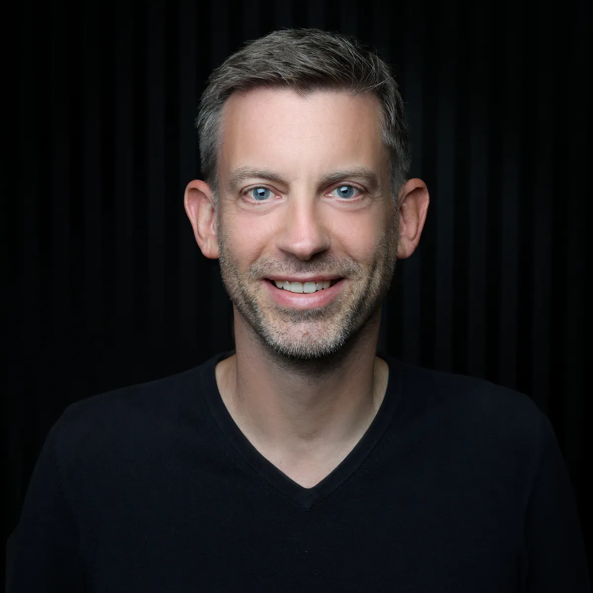 Sebastian Wolter in a black V-neck jumper against a dark background, smiling at the camera