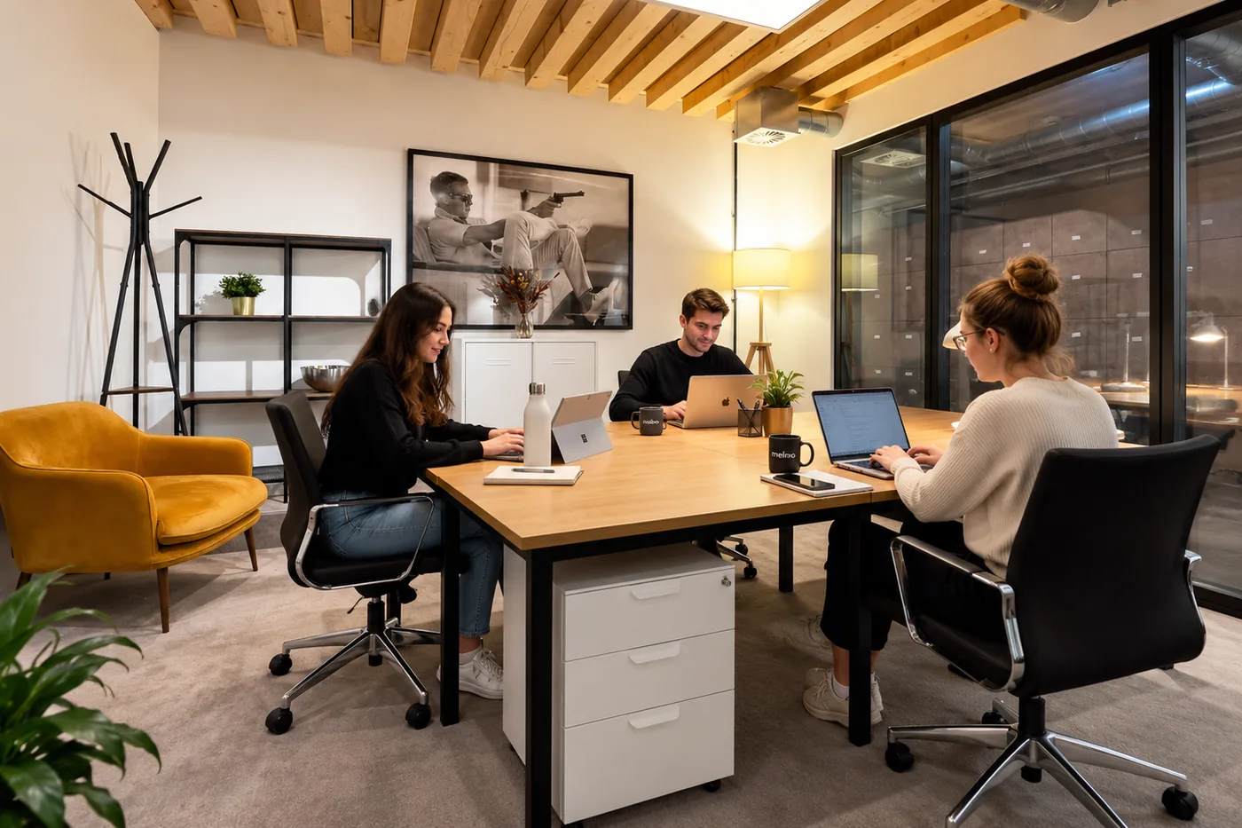 Three colleagues at work in one of the melino team rooms, laptops open around a shared desk under a beamed ceiling, with the office visible through the glazed wall behind