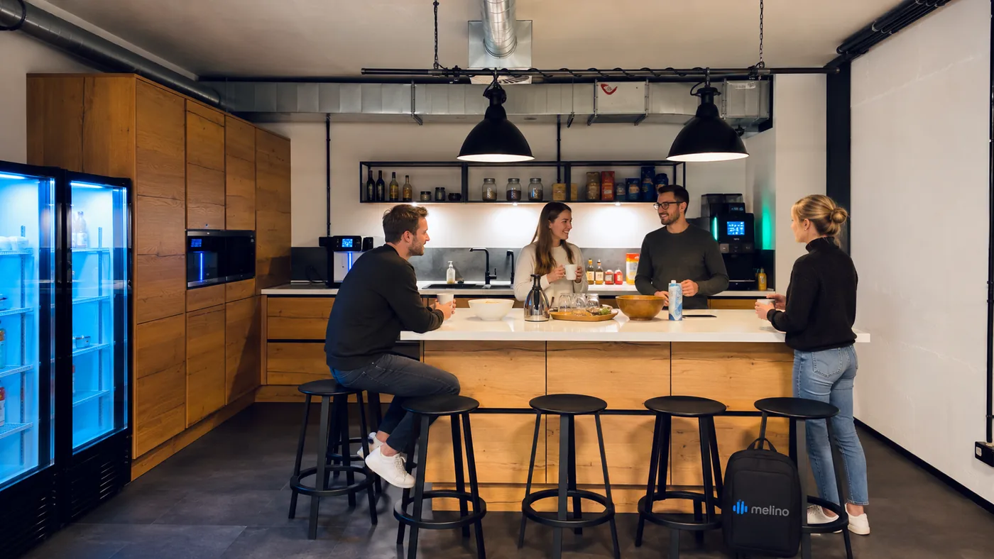 Four colleagues standing and sitting around the office kitchen island with coffee, under two black pendant lamps, a coffee machine and a stocked fridge behind them